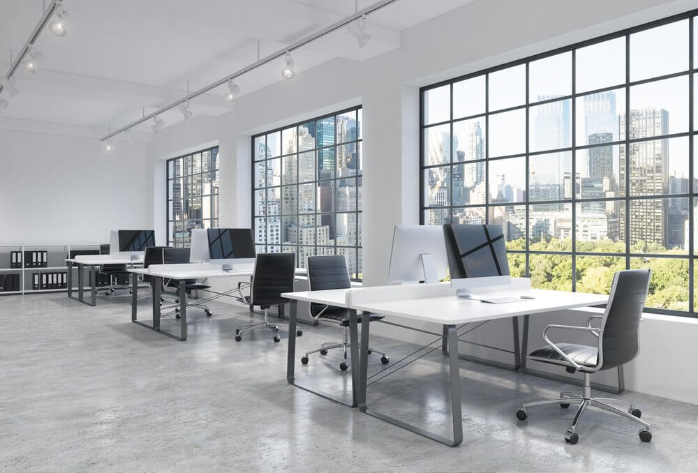 An open-plan office space with large windows, ergonomic chairs, and standing desks, overlooking a city skyline.